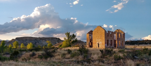 Ruins of a stone building surrounded by grass and trees, with dramatic clouds in the sky and hills in the background.