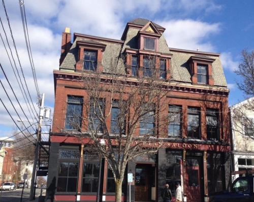 Historic brick building with a sloped roof, large windows, and a sign reading "The Saloon" under a partly cloudy sky.