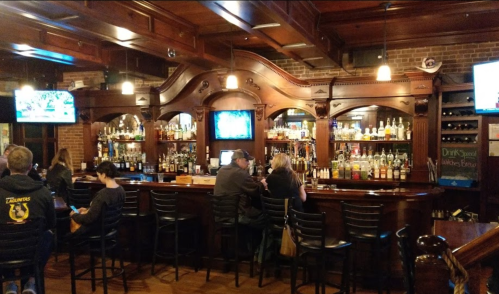 A cozy bar with wooden decor, shelves of liquor, and patrons seated at the counter, watching TVs.
