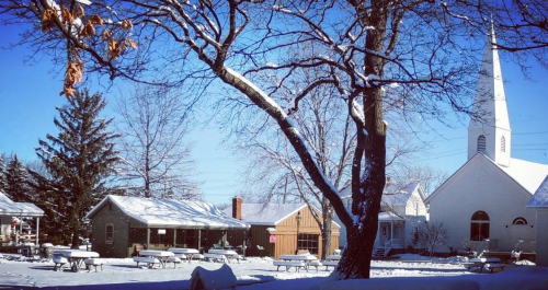 A snowy landscape featuring a church, houses, and bare trees under a clear blue sky. Picnic tables are visible in the foreground.
