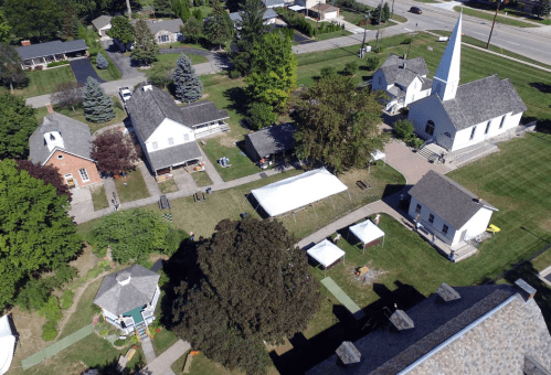 Aerial view of a small village with historic buildings, a church, and tents set up on green lawns.