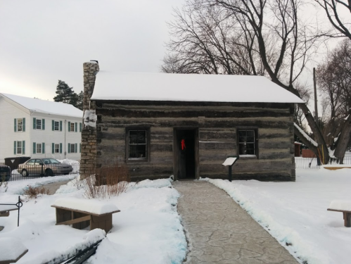 A log cabin covered in snow, with a stone chimney and a path leading to the door, surrounded by winter scenery.