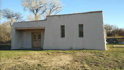 A simple, white adobe building with a flat roof, surrounded by grass and trees, under a clear blue sky.