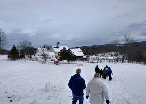A group of people walking through snow towards a farm with a church steeple and mountains in the background.