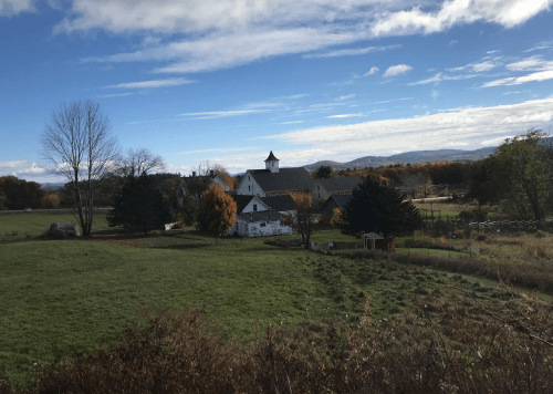 A scenic view of a farm with a white barn, surrounded by trees and mountains under a blue sky with clouds.