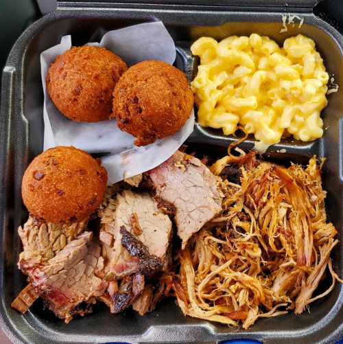 A takeout container with sliced brisket, pulled pork, macaroni and cheese, and three cornbread muffins.