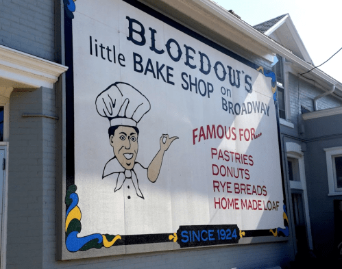 Sign for Bloedow's Bake Shop on Broadway, featuring a chef and listing pastries, donuts, rye breads, and homemade loaf.
