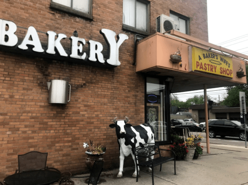 A bakery storefront featuring a sign, a cow statue, and outdoor seating with flowers.