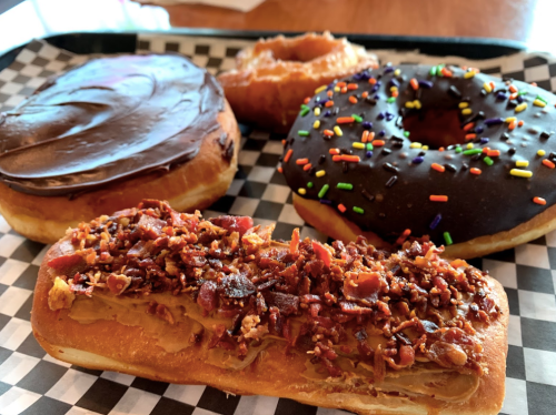 A tray of four donuts: a chocolate frosted, a glazed, a chocolate with sprinkles, and a bacon-topped pastry.