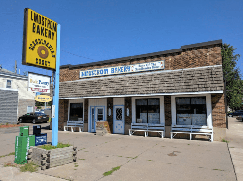 A brick bakery with a sign reading "Lindstrom Bakery" and "Scandinavian Donut," featuring a sunny blue sky.