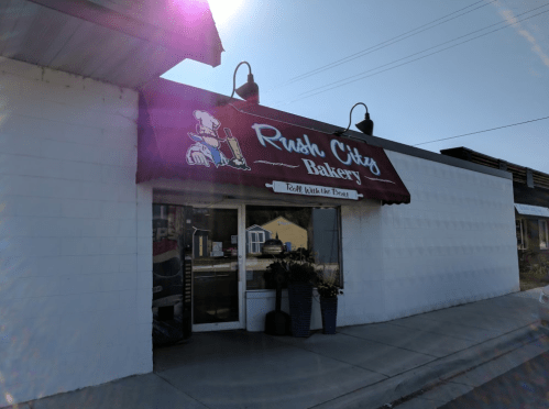 Exterior of Rush City Bakery with a red awning and a sign featuring a baker, surrounded by potted plants.