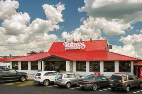 A red-roofed restaurant with a sign reading "Tobie's Restaurant," surrounded by parked cars and a cloudy sky.
