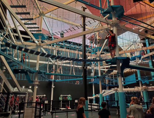 A vibrant indoor ropes course with people navigating obstacles high above, surrounded by colorful structures.