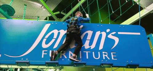 A person in a harness climbs a large blue sign for Jordan's Furniture indoors.