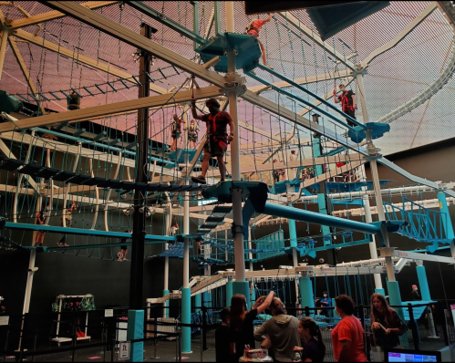A vibrant indoor ropes course with people climbing and navigating obstacles, surrounded by spectators below.