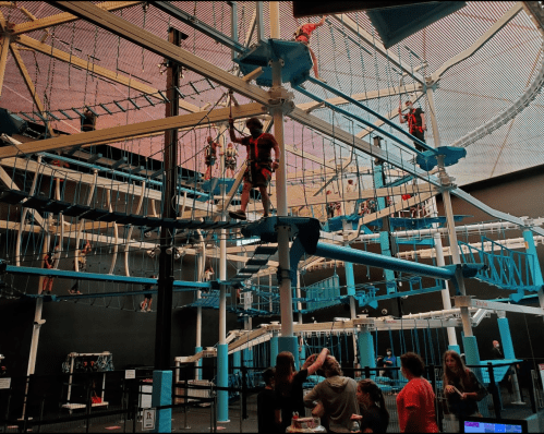 A vibrant indoor ropes course with people climbing and navigating obstacles, surrounded by spectators below.