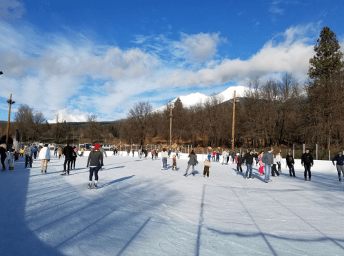 A busy outdoor ice skating rink with people skating under a blue sky and mountains in the background.