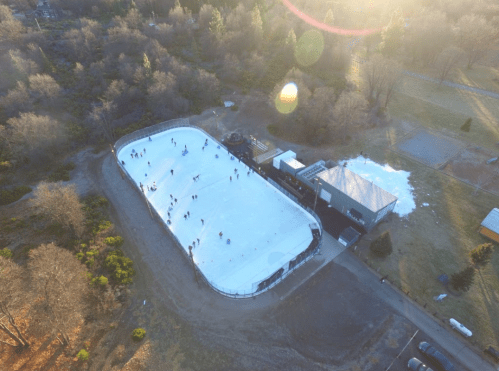 Aerial view of an outdoor ice skating rink with people skating, surrounded by trees and a building nearby.
