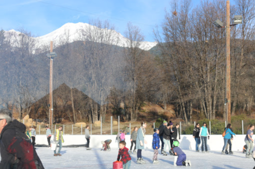 A busy outdoor ice skating rink with people of all ages skating and enjoying the winter scenery and mountains in the background.
