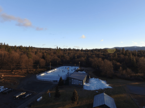Aerial view of an outdoor ice rink surrounded by trees and mountains, with skaters enjoying the sunny day.