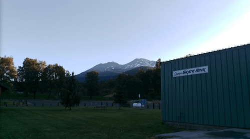 A skate rink building with a mountain backdrop and trees under a clear sky.