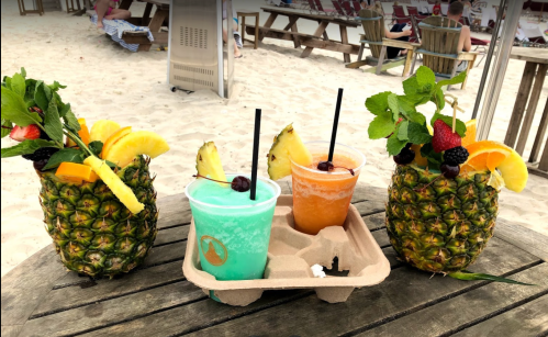 Three tropical drinks served in pineapples and cups, garnished with fruit, on a wooden table at a beach.