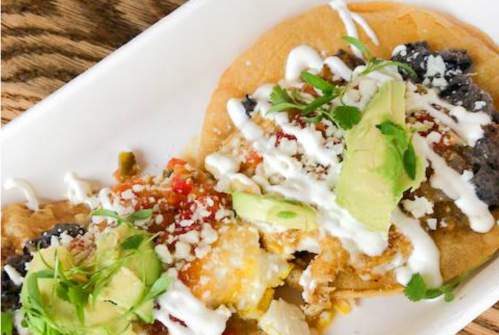 Three colorful tacos topped with avocado, cheese, sour cream, and fresh herbs on a white plate.