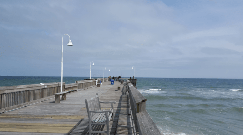 A wooden pier extends over the ocean, with people in the distance and cloudy skies above.