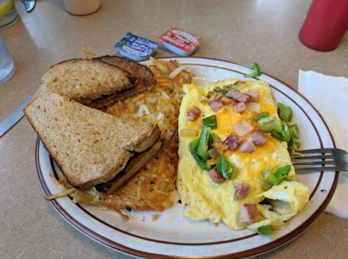 A plate with a ham and green pepper omelet, hash browns, and two slices of toasted bread with condiments on the side.