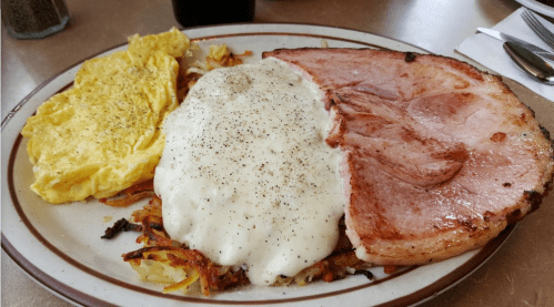 A plate of breakfast featuring an omelet, hash browns topped with gravy, and a slice of ham.