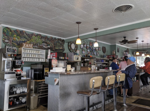 A cozy café interior with a counter, coffee machine, and patrons seated at the bar, surrounded by colorful wall art.