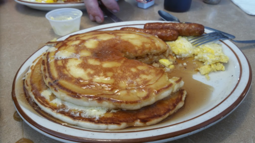 A plate of fluffy pancakes with syrup, scrambled eggs, and sausage links on a diner table.