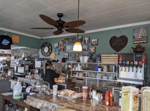 A cozy café interior with a counter, a woman preparing food, and walls adorned with photos and decorations.