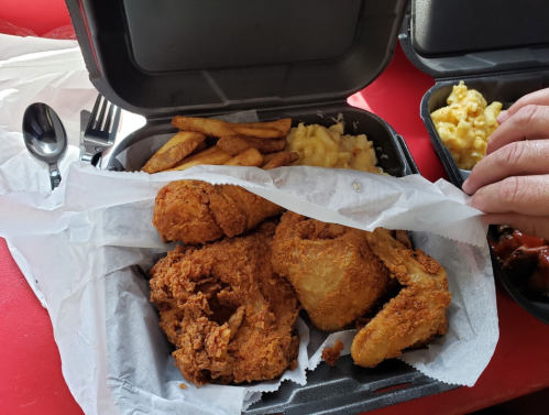 A takeout container with fried chicken, fries, coleslaw, and macaroni and cheese, with a hand lifting the lid.