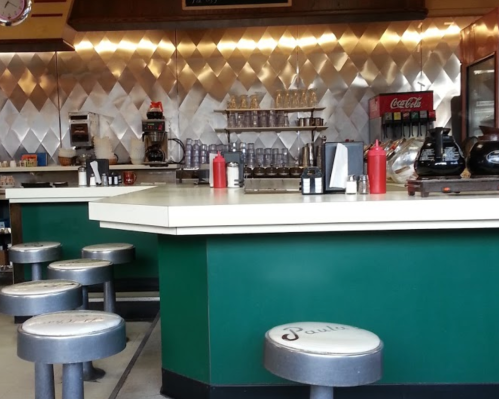 A retro diner interior featuring a counter, stools, and a shiny metallic backsplash with condiments on the counter.