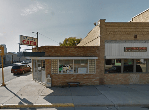 Exterior view of Paula's Cafe, a brick building with a sign, located on a street corner. Cars parked nearby.