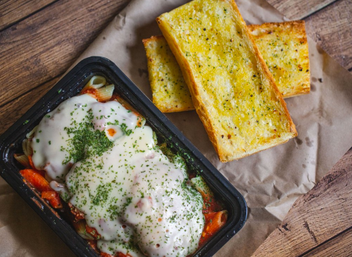 A black tray with cheesy pasta topped with herbs, alongside two pieces of golden garlic bread on brown paper.