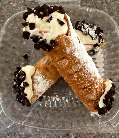 Three cannoli filled with cream, topped with chocolate chips and powdered sugar, in a clear plastic container.