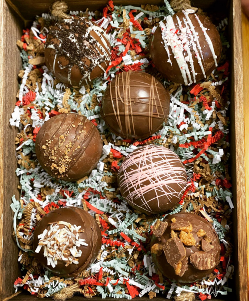 A wooden tray filled with colorful shredded paper holds seven decorative chocolate truffles, each with unique toppings.
