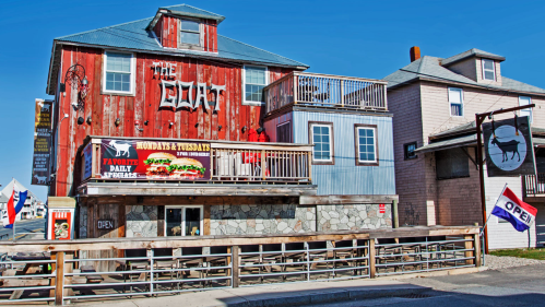 A rustic restaurant with a red wooden exterior, balconies, and a sign reading "The Goat," featuring outdoor seating.