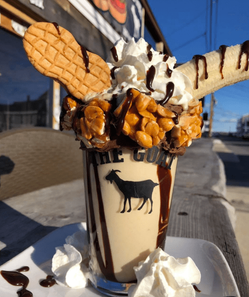 A decadent milkshake topped with whipped cream, peanuts, cookies, and chocolate drizzle, served in a glass with a goat logo.