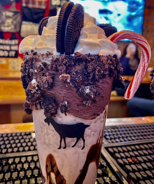 A decadent milkshake topped with whipped cream, crushed Oreos, and a candy cane, served in a glass with a cow logo.