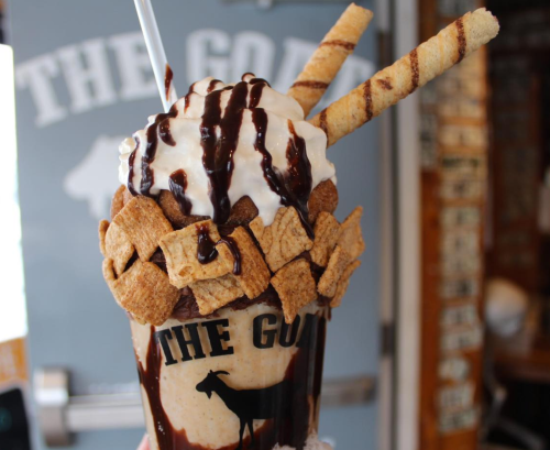 A decadent milkshake topped with whipped cream, chocolate drizzle, cereal, and wafer sticks, served in a branded glass.