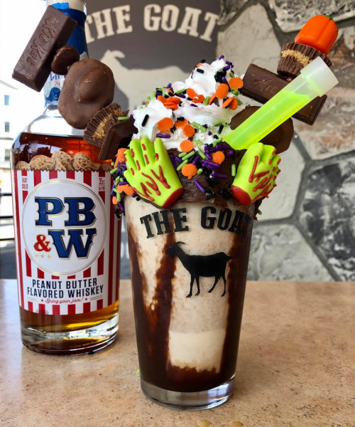 A decadent milkshake topped with whipped cream, candy, and a shot of peanut butter whiskey in the background.