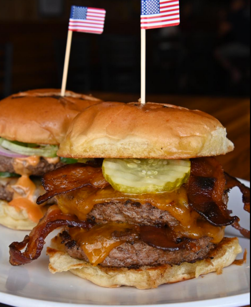 A close-up of two burgers with bacon, cheese, pickles, and small American flags on top, served on a white plate.