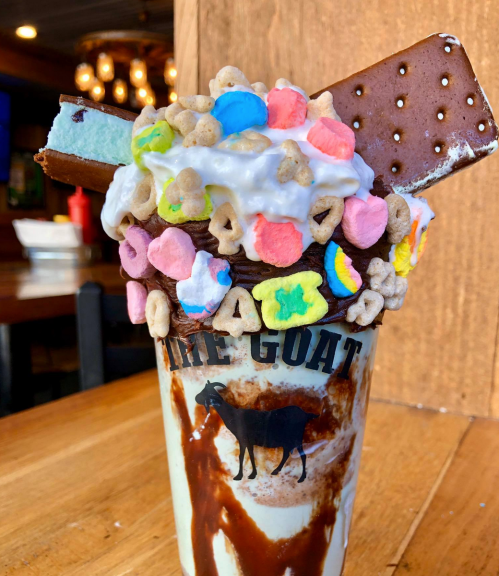 A colorful dessert in a cup topped with whipped cream, cereal, and ice cream sandwiches, with a chocolate drizzle.