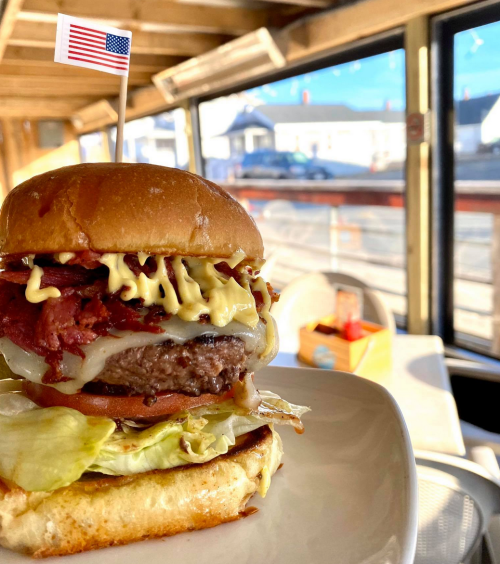 A close-up of a delicious burger with lettuce, cheese, and a flag, set against a bright, sunny restaurant interior.