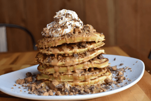 A tall stack of pancakes topped with whipped cream, peanut butter drizzle, and crushed chocolate pieces on a white plate.
