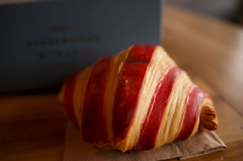 A beautifully crafted croissant with red stripes, resting on a brown paper napkin, with a blue box in the background.