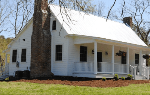 A white, single-story house with a metal roof, brick chimney, and a front porch surrounded by green grass and landscaping.
